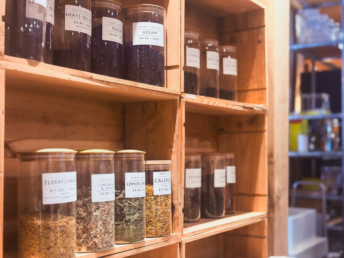 Wooden shelves stocked with clear glass jars containing various dried herbs and spices, each with a white paper label showing the name of the ingredient.