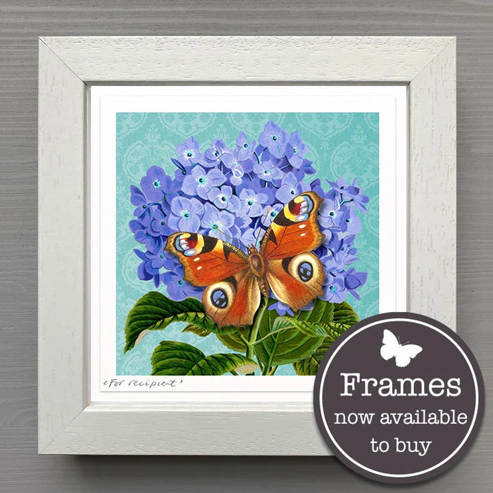 A framed botanical art print featuring purple hydrangeas and a butterfly on green leaves, set in a white textured frame against a gray wall. A circular badge in the lower right says 'Frames now available to buy'.