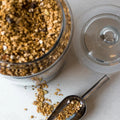 Glass container with granola, metal scoop, and scattered granola on a light surface