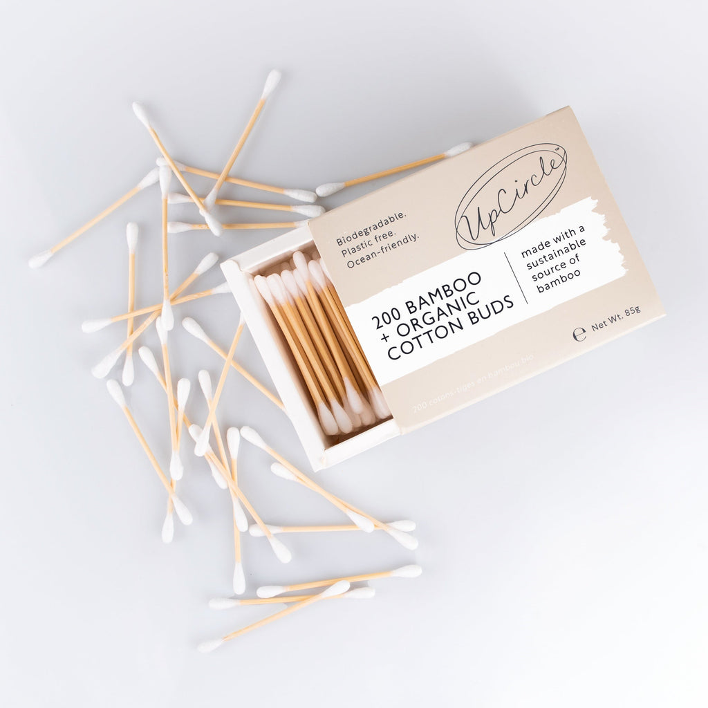 An open box of cotton buds with several loose cotton swabs scattered around it on a light gray background.