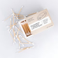 An open box of cotton buds with several loose cotton swabs scattered around it on a light gray background.