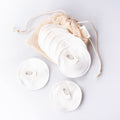 Three white reusable make-up remover pads with small oval brand tags, arranged with a beige drawstring mesh storage bag on a clean white background.