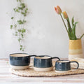 Three ceramic mugs with dark navy blue and cream glaze are arranged on a woven placemat on a wooden table, with a vase of tulips in the background.
