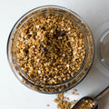 Granola in a clear glass jar with a metal scoop beside it, photographed from above on a light neutral background.