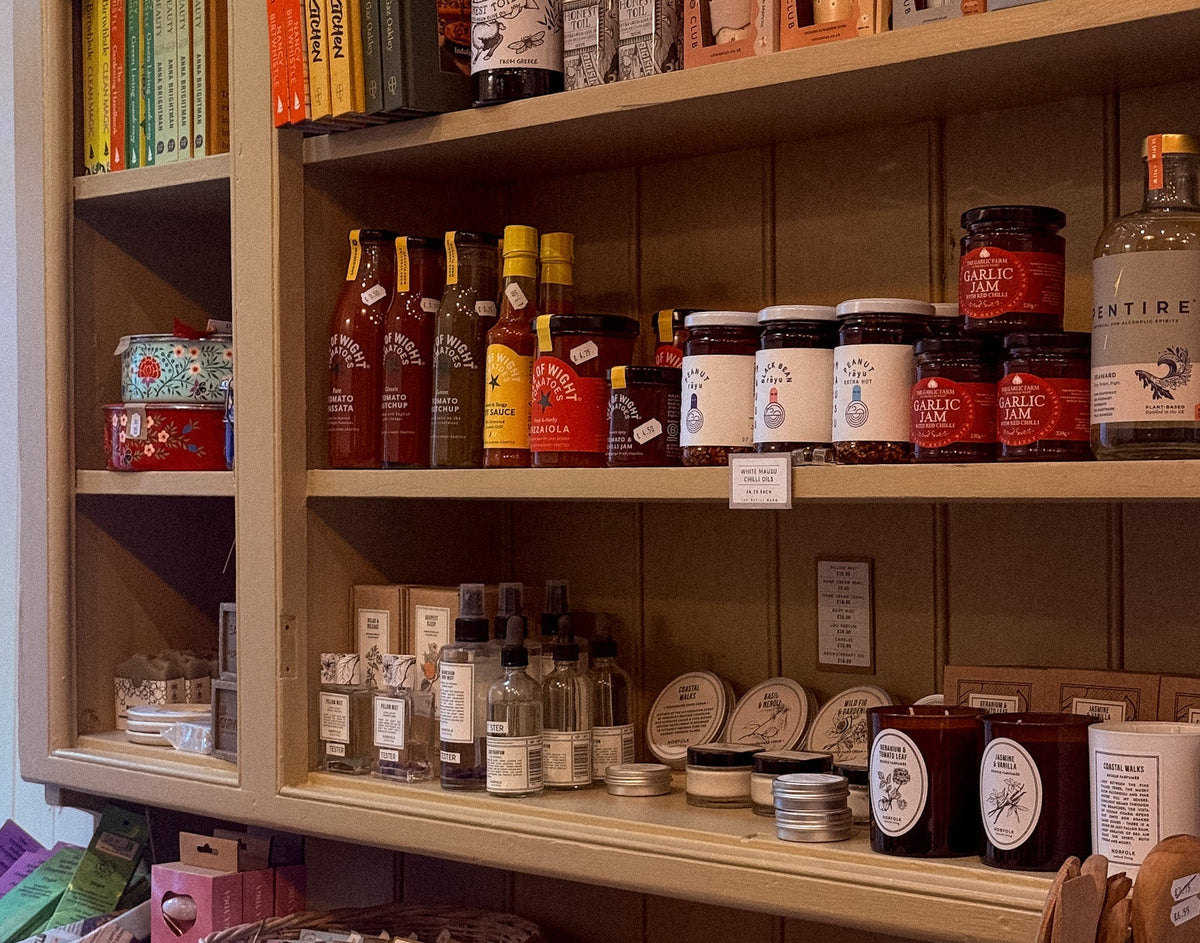 A wooden display shelf in a shop filled with assorted home goods, jars, bottles, books, baskets, and packaged items, photographed in a real retail environment.