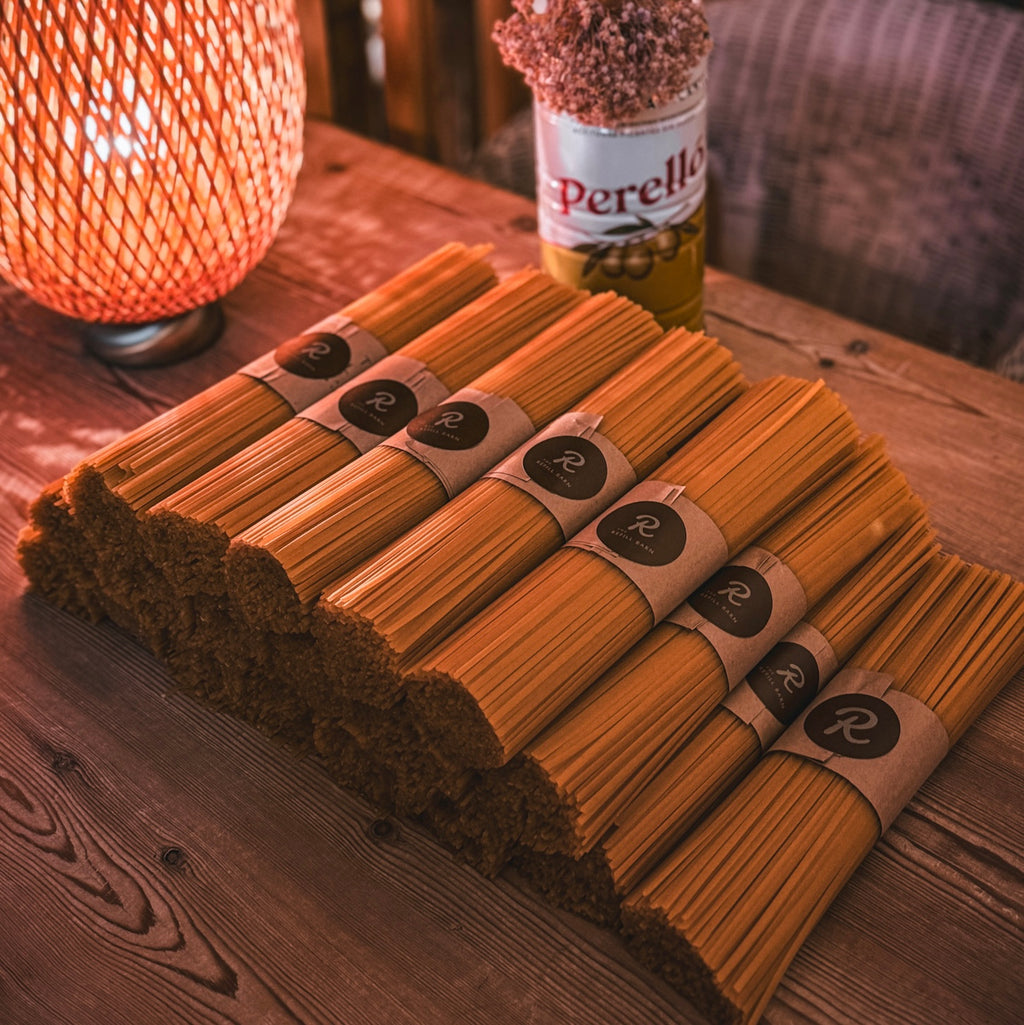 Several bundles of dried linguine pasta tied with paper bands are arranged on a wooden table, with a warm glowing lamp and a bottle in the background.