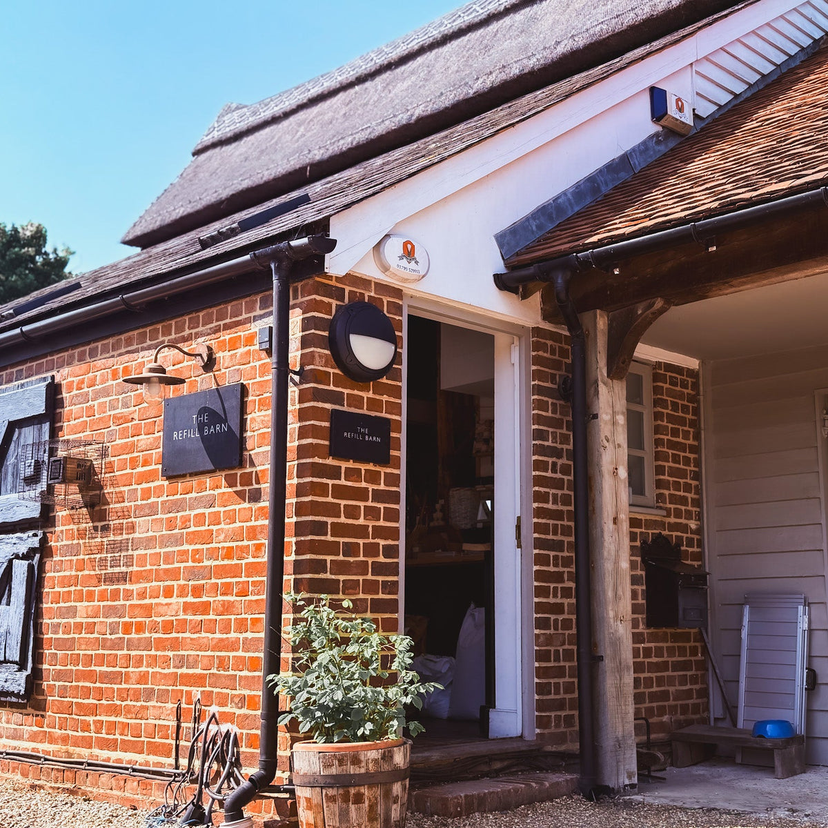 A rustic brick cottage with a thatched roof, open doorway, wooden support beams, and a white garage door under a tiled roof. A small sign on the brick wall appears to read a brand name, and the scene is photographed in bright daylight with plants in the foreground.