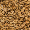Golden-brown baked granola in a dark gray baking pan lined with parchment paper, with a black and white checkered cloth visible in the top left corner.