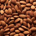 This close-up photograph displays a pile of almonds on a dark surface. The almonds are tightly packed together, creating a textured pattern. The brown hues of the almonds contrast with the black background, highlighting the natural colors and shapes of the nuts.
