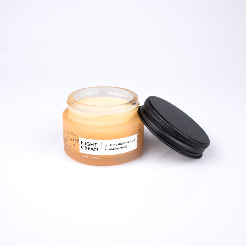 A small clear glass jar of UpCircle night cream with a black screw-on lid resting beside it on a clean white background.