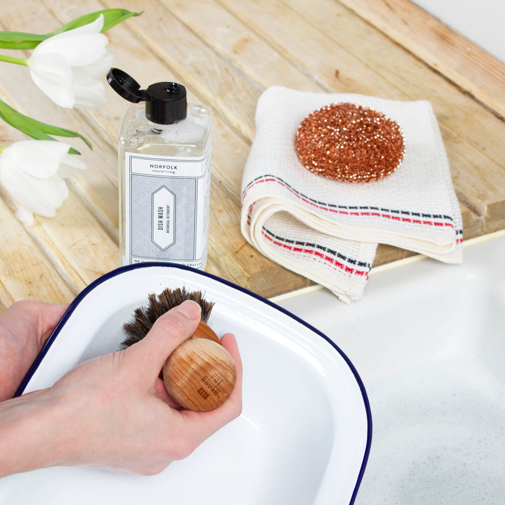 Person cleaning a dish with a brush, next to a bottle of soap and a towel on a wooden surface.