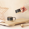 A hand holds a dark glazed ceramic mug above another matching mug resting on a light wood board, with a warm neutral background and decorative star-shaped lights.