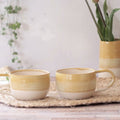 Two ceramic mugs with a yellow and cream horizontal striped design, sitting on a woven placemat on a wooden table with a blurred background.