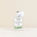 A clear glass perfume bottle labeled 'Pillow Mist' with a white floral cap insert and a green crystalline scent element at the bottom, set against a warm beige background.