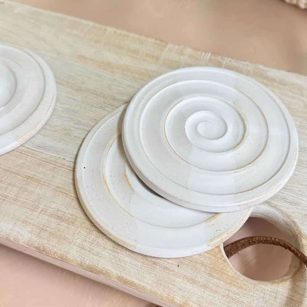 Three round ceramic coasters with a raised spiral design, featuring a glossy off-white glaze and subtle gold speckles, arranged on a light wood serving board.