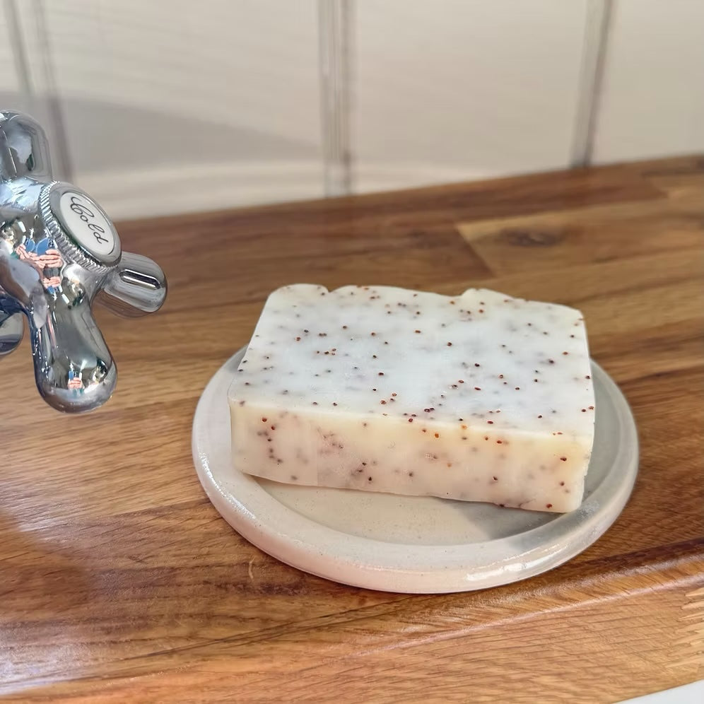 A rectangular bar of handmade soap with white and beige marbling and small brown specks, resting on a small round ceramic dish on a wooden countertop next to a chrome faucet.